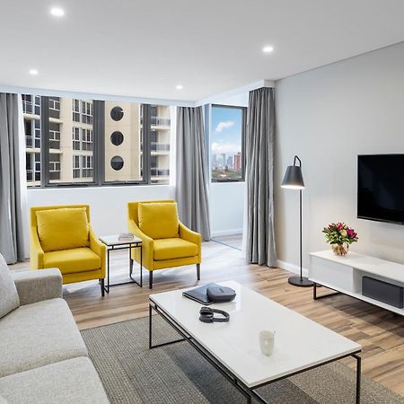 Meriton Suites Bondi Junction Сідней Екстер'єр фото The photo shows a modern, well-lit living room. It features a light gray sofa with a textured fabric. There are two bright yellow armchairs positioned near a window, which allows natural light to enter the space. The room has light gray curtains and 
