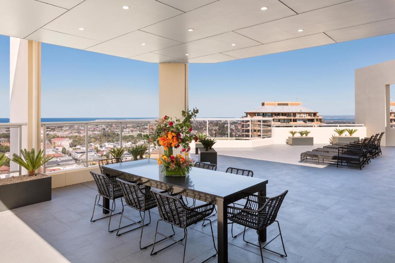 Meriton Suites Bondi Junction Сідней Екстер'єр фото The photo shows a stylish outdoor terrace or balcony area, featuring a long dining table surrounded by several black wire chairs. The table has a centerpiece of vibrant flowers. The space is well-lit, with a clear view of the horizon in the backgroun