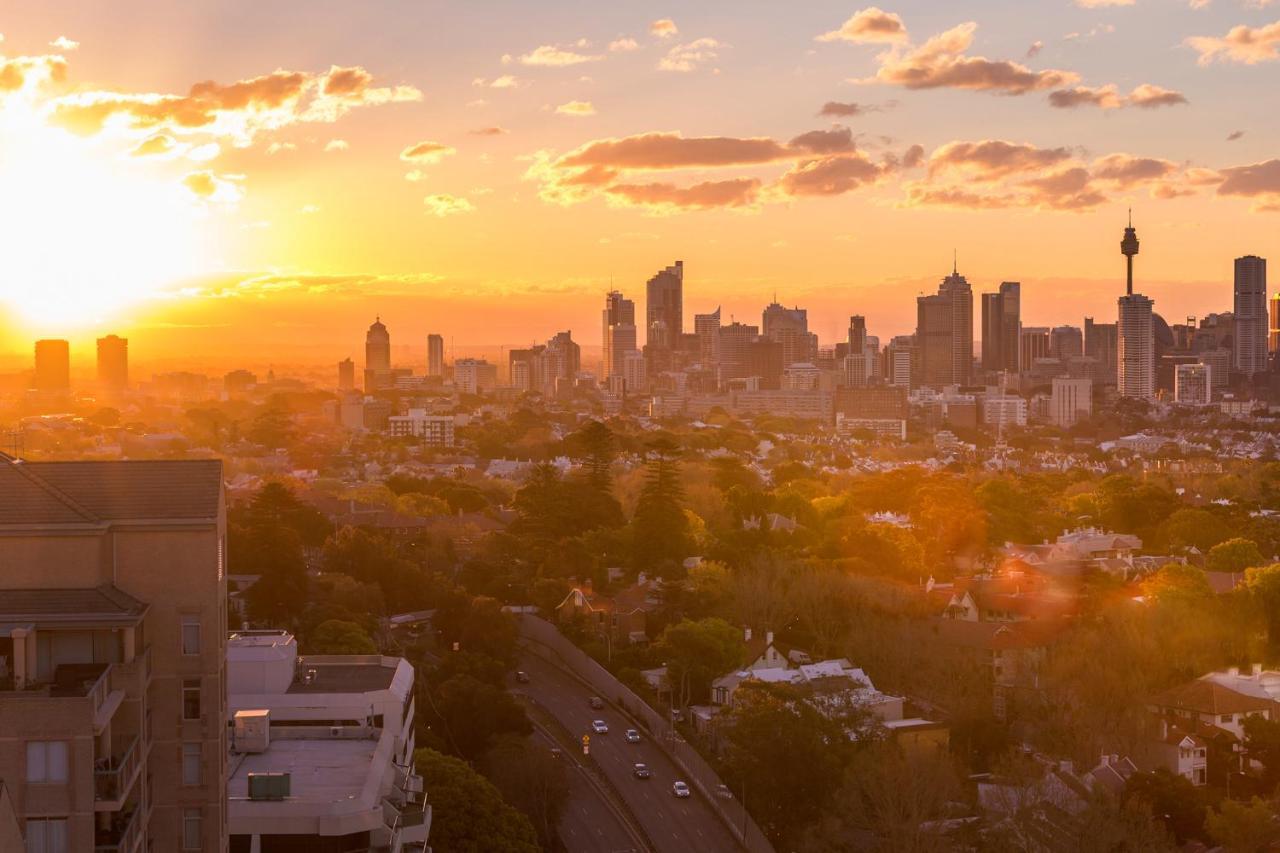 Meriton Suites Bondi Junction Сідней Екстер'єр фото The photo shows a panoramic view of a city skyline at sunset. The sun is low on the horizon, casting a warm golden light across the buildings. Tall skyscrapers are silhouetted against the colorful sky, which features hues of orange, pink, and yellow,
