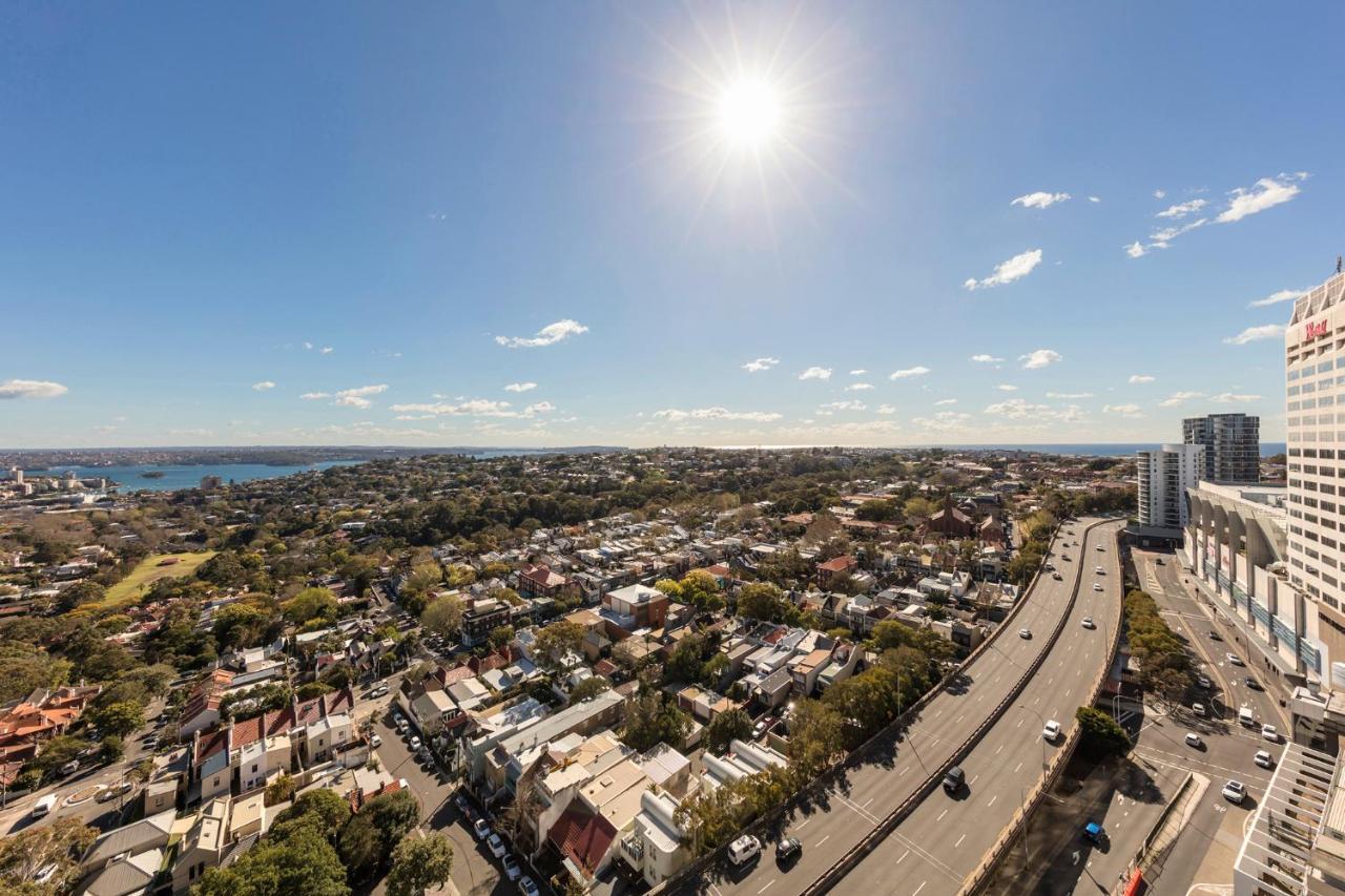 Meriton Suites Bondi Junction Сідней Екстер'єр фото The photo shows a panoramic view of a city landscape under a clear blue sky. In the foreground, there is a road with vehicles traveling along it, bordered by trees and buildings. In the background, you can see a residential area with various houses, 