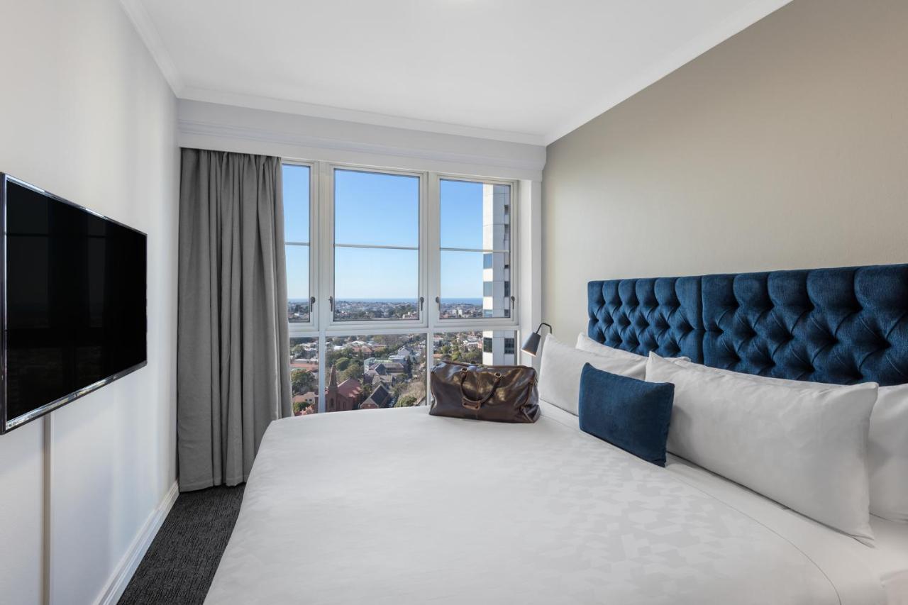 Meriton Suites Bondi Junction Сідней Екстер'єр фото The photo shows a modern hotel room featuring a large bed with a tufted blue headboard. Beside the bed, there is a stylish bag placed on the bed. The room has large windows that provide a view of the outside, allowing natural light to fill the space.