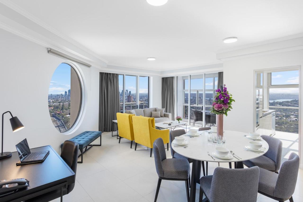 Meriton Suites Bondi Junction Сідней Екстер'єр фото The photo shows a modern and bright interior space, likely a living room or dining area. It features large windows that offer a panoramic view of the city skyline. The furnishings include a round dining table set for several people, with plates and g
