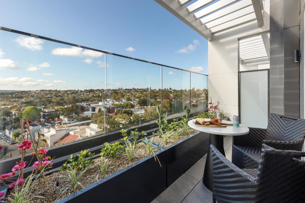 Meriton Suites Bondi Junction Сідней Екстер'єр фото The photo shows a modern balcony with a glass railing, enhancing the view of the surrounding area, which includes various rooftops and greenery in the distance. There is a small round table set with a decorative floral arrangement and some items, lik