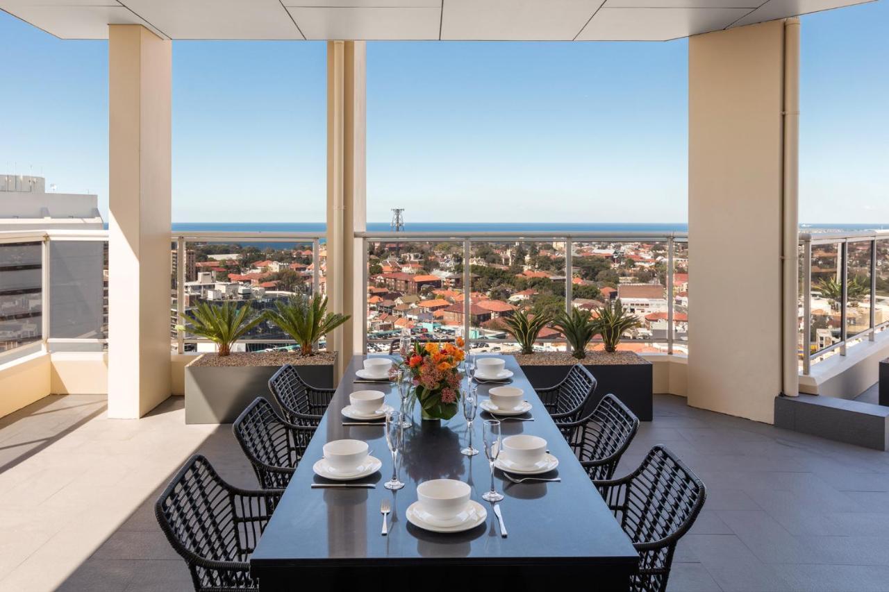 Meriton Suites Bondi Junction Сідней Екстер'єр фото The photo shows a modern outdoor dining area situated on a balcony or terrace with a stunning view. There is a long rectangular table set with plates, glasses, and cutlery, ready for a meal. The table is surrounded by sleek, black wicker chairs. In t
