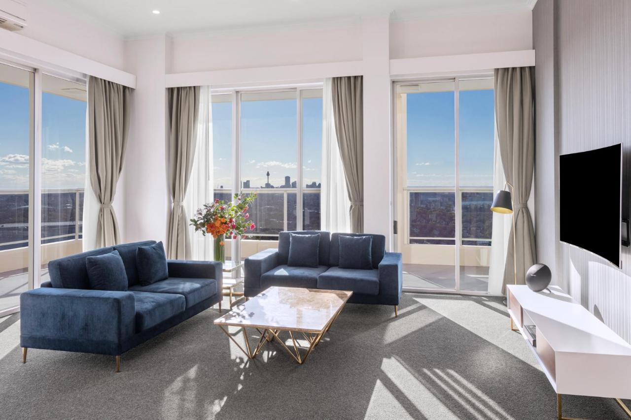 Meriton Suites Bondi Junction Сідней Екстер'єр фото The photo shows a modern living room with large windows that allow natural light to fill the space. There are two blue sofas arranged around a coffee table with a glass top and gold legs. A floral arrangement adds a touch of color, placed on a nearby
