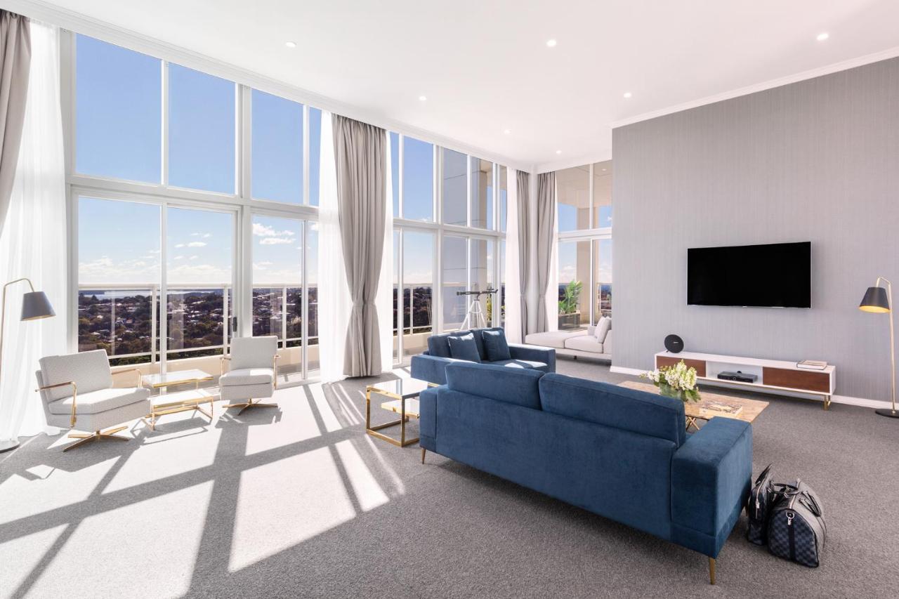 Meriton Suites Bondi Junction Сідней Екстер'єр фото The photo shows a bright and modern living room with large floor-to-ceiling windows that provide a view of the outdoors. The space features a blue sofa in the center, complemented by light-colored armchairs nearby. There are light curtains hanging by