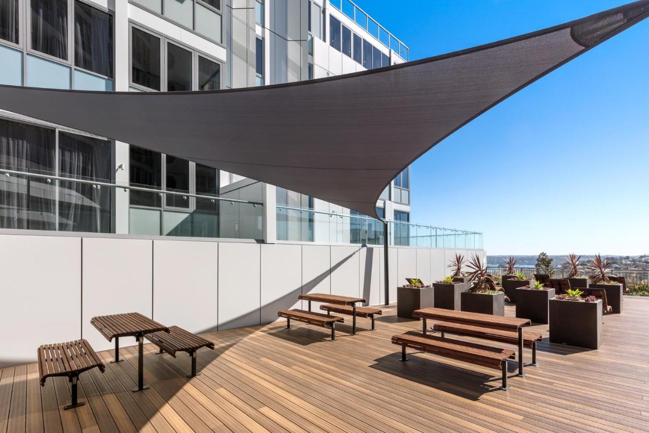 Meriton Suites Bondi Junction Сідней Екстер'єр фото The photo shows a modern outdoor terrace area on a building. It features several wooden benches arranged on a wooden deck. There are large planters with greenery, adding some decorative elements. A large triangular gray shade sail is positioned above