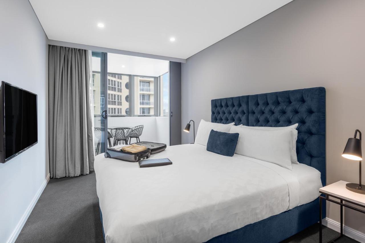Meriton Suites Bondi Junction Сідней Екстер'єр фото The photo shows a modern hotel room featuring a large bed with a tufted blue headboard. There are minimalistic bedside tables with lamps on either side of the bed. A flat-screen TV is mounted on the wall across from the bed. To one side, there are fl