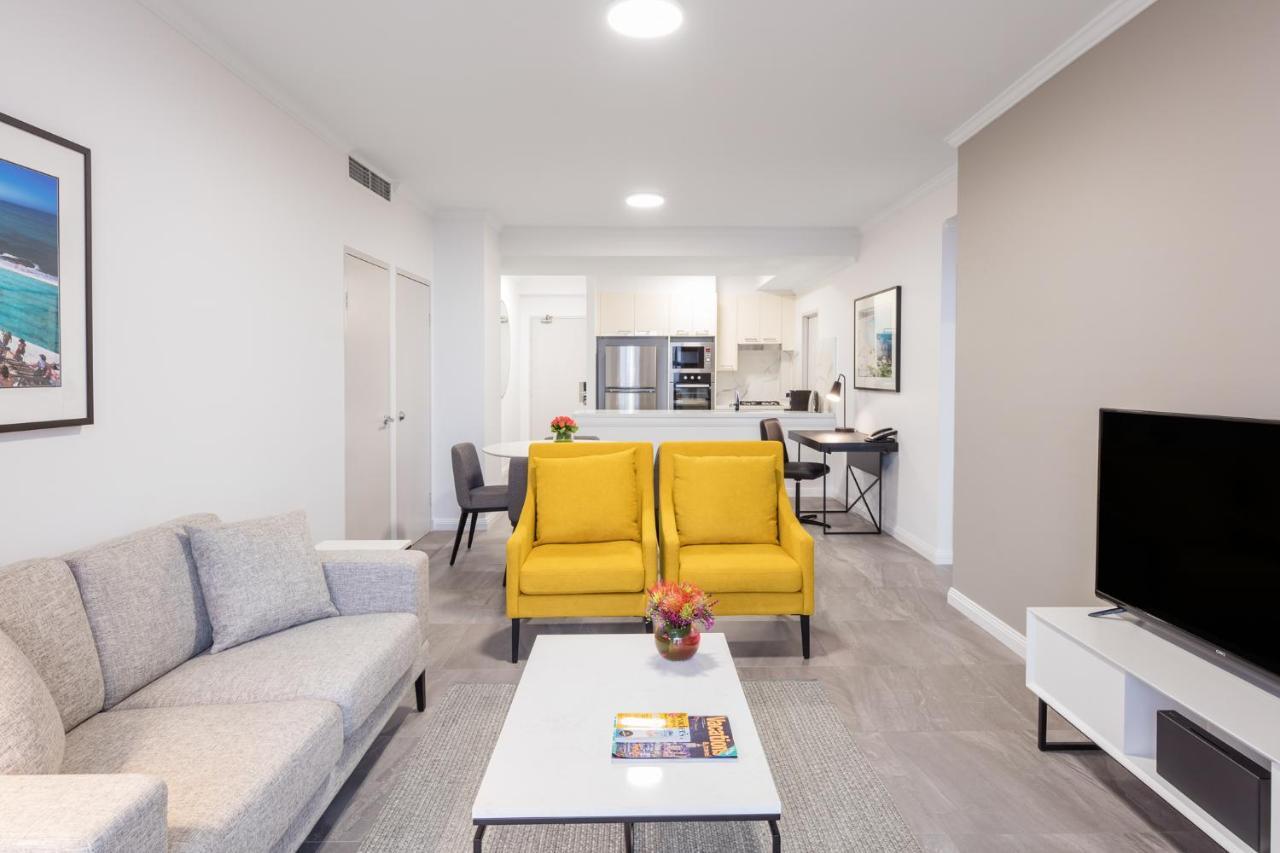 Meriton Suites Bondi Junction Сідней Екстер'єр фото The photo shows a modern living space with a light and airy design. In the foreground, there are two bright yellow armchairs flanking a white coffee table, which has a small vase of flowers and a magazine on it. To the left, there is a light gray sof