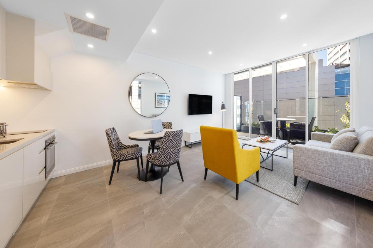 Meriton Suites Bondi Junction Сідней Екстер'єр фото The photo shows a modern and stylish living space. On one side, there is a kitchen area with sleek cabinetry, and next to it, a round dining table surrounded by three patterned chairs. In the center of the room, there is a gray sofa accompanied by a 