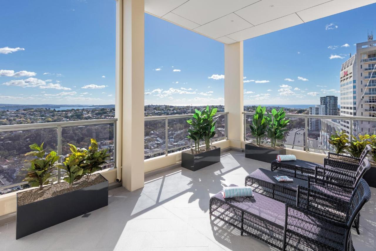 Meriton Suites Bondi Junction Сідней Екстер'єр фото The photo shows a modern balcony or terrace area with a stunning view. There are several planters with green plants placed along the edge. The seating area features lounge chairs in a contemporary design, likely made of woven materials. The space app