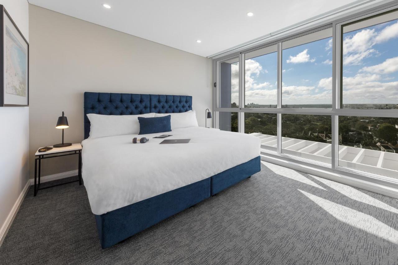 Meriton Suites Bondi Junction Сідней Екстер'єр фото The photo shows a modern and stylish hotel room. It features a comfortable queen-size bed with a blue tufted headboard. The bed is neatly made with white linens and pillows. Next to the bed, there is a small bedside table with a desk lamp. 

On the f