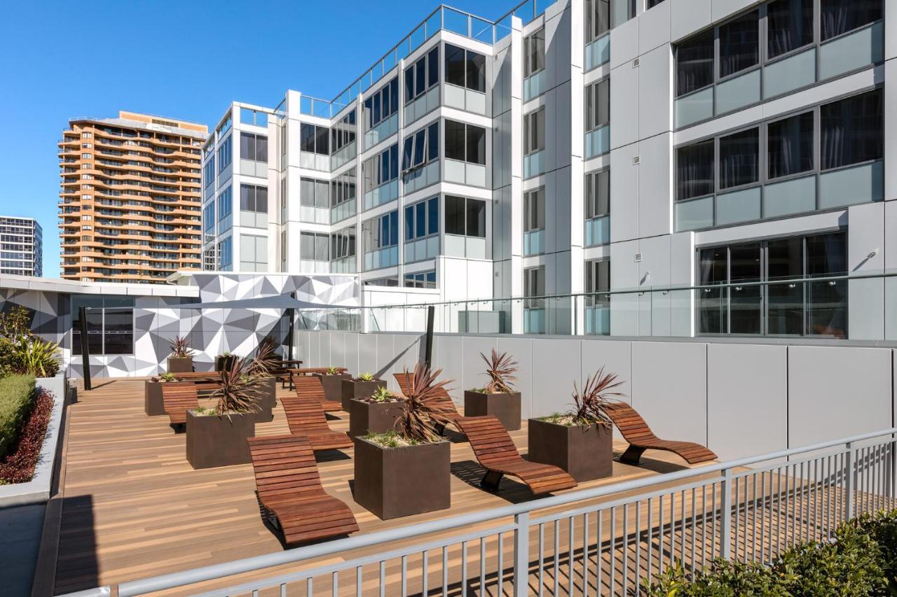 Meriton Suites Bondi Junction Сідней Екстер'єр фото The photo shows a modern outdoor space featuring a wooden deck. There are several lounge chairs arranged around large planter boxes, which contain plants. In the background, there are contemporary residential buildings with large windows, and a geome