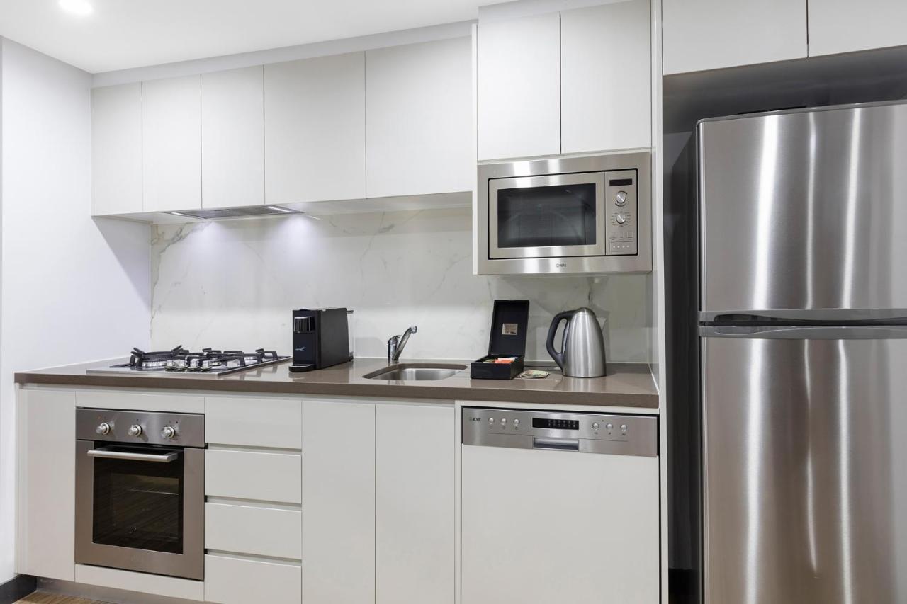 Meriton Suites Bondi Junction Сідней Екстер'єр фото The photo shows a modern kitchen with a sleek, minimalist design. It features white cabinetry and a countertop that blends with the overall aesthetic. 

Key elements include:

- A stainless steel refrigerator on the right.
- An oven with a gas cookto