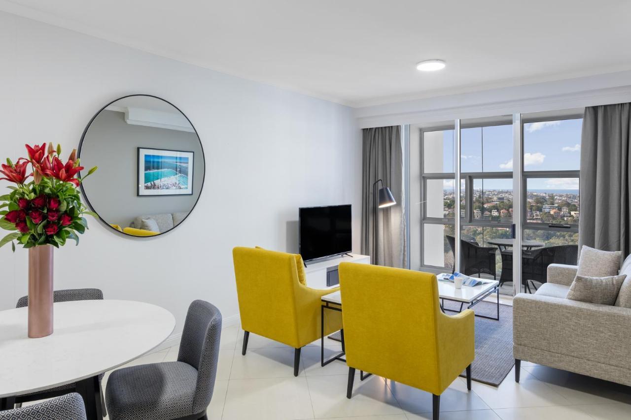 Meriton Suites Bondi Junction Сідней Екстер'єр фото The photo shows a modern living space with a stylish design. There are two bright yellow chairs facing a small glass table, and a comfortable beige sofa to the right. A TV is positioned on the wall in front of the chairs. To the left, there's a dinin