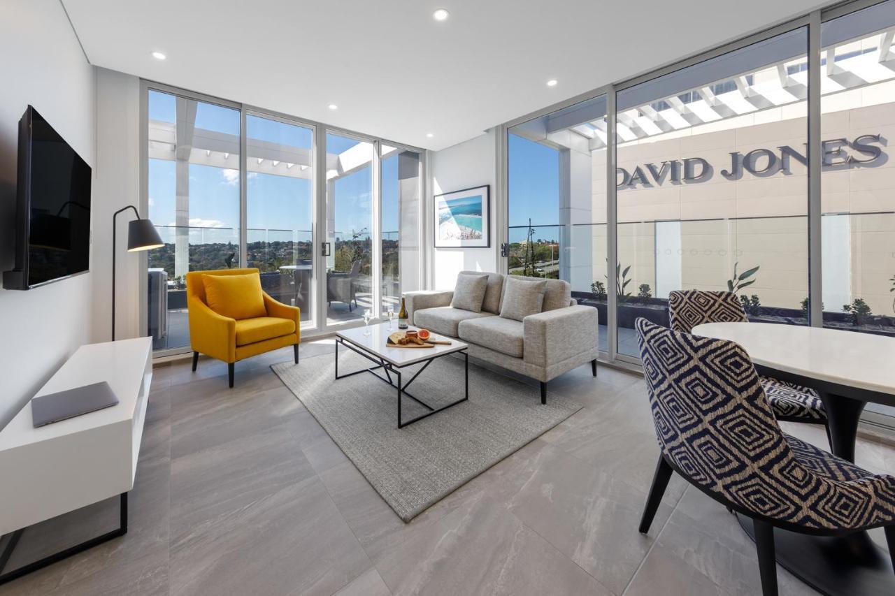 Meriton Suites Bondi Junction Сідней Екстер'єр фото The photo shows a modern living space that features large windows allowing ample natural light. The room is furnished with a gray sofa and a bright yellow armchair, along with a coffee table in between. There’s a decorative bowl with fruits on the ta