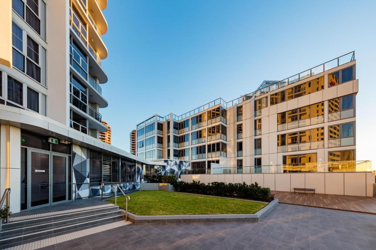 Meriton Suites Bondi Junction Сідней Екстер'єр фото The photo showcases a modern residential area featuring contemporary buildings. In the foreground, there is a landscaped green space surrounded by well-designed architecture. The buildings have large glass windows, reflecting the light, and feature a