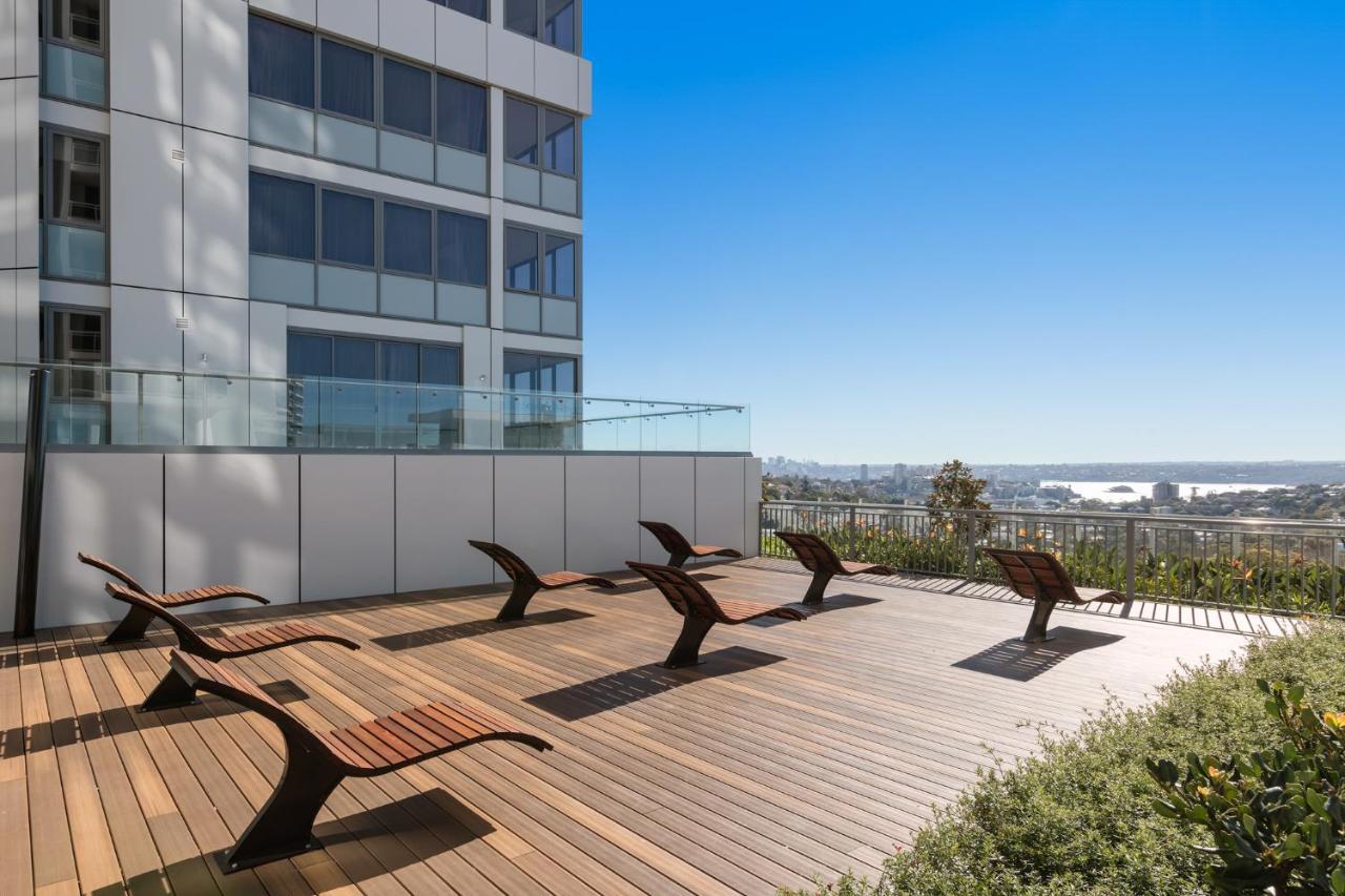 Meriton Suites Bondi Junction Сідней Екстер'єр фото The photo shows a rooftop or outdoor area with a wooden deck. There are several modern lounge chairs arranged neatly, facing a clear sky and a panoramic view of the surrounding landscape. In the background, there is a tall building with large windows