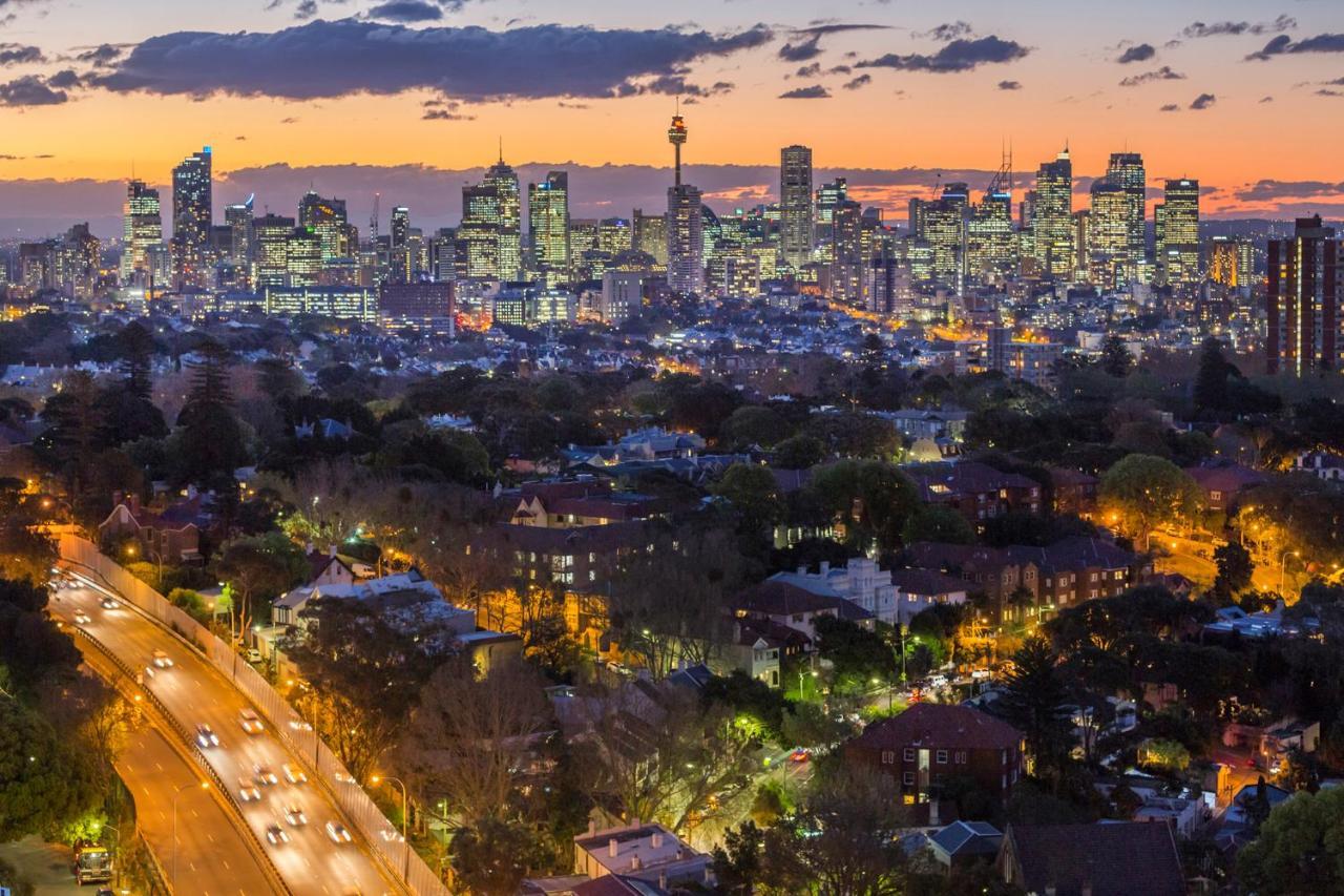 Meriton Suites Bondi Junction Сідней Екстер'єр фото The photo captures a stunning city skyline at dusk, featuring towering skyscrapers adorned with lights against a colorful sunset backdrop. In the foreground, a green area with trees and houses is visible, while a road with cars can be seen winding th