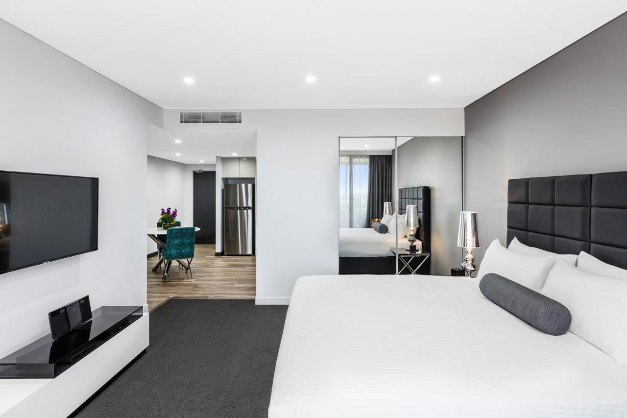 Meriton Suites Bondi Junction Сідней Екстер'єр фото The photo shows a modern hotel room. It features a large bed with a white covering and gray accent pillows. To the left, there's a flat-screen TV mounted on the wall above a sleek white cabinet. The flooring is a light wood tone, and a large mirror i