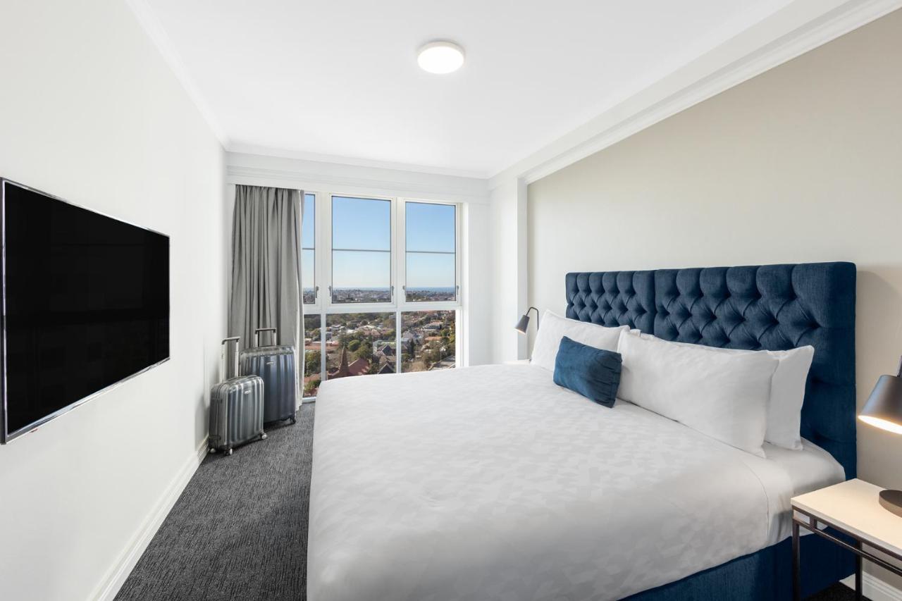 Meriton Suites Bondi Junction Сідней Екстер'єр фото The photo shows a modern hotel room with a large bed featuring a tufted blue headboard. There is a white duvet and a decorative pillow on the bed. A flat-screen television is mounted on the wall opposite the bed. Large windows are covered with gray c