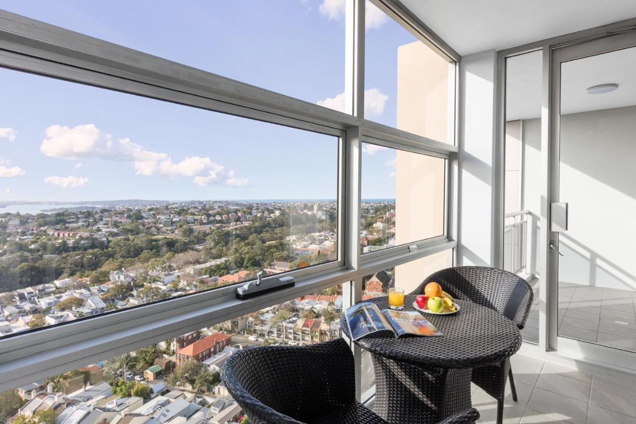 Meriton Suites Bondi Junction Сідней Екстер'єр фото The photo shows a modern balcony with large glass windows that offer a panoramic view of a sprawling urban landscape. There is a small round table made of dark wicker, accompanied by two matching chairs. On the table, there is a bowl of fruit and a m