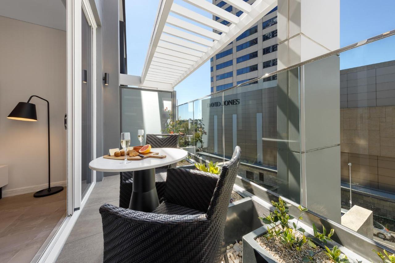 Meriton Suites Bondi Junction Сідней Екстер'єр фото The photo shows a modern balcony area with glass railings. It features a small round table set for two, with a plate of food and drinks. There are two stylish wicker chairs positioned around the table. The space is bright and airy, with sunlight stre