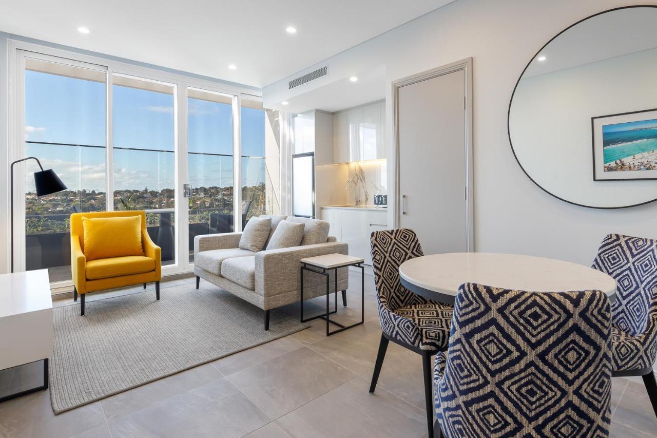 Meriton Suites Bondi Junction Сідней Екстер'єр фото The photo shows a modern and stylish living area. It features a comfortable sofa in a neutral color, accompanied by a bright yellow armchair that adds a pop of color. There's a round dining table with patterned chairs nearby. Large windows allow plen