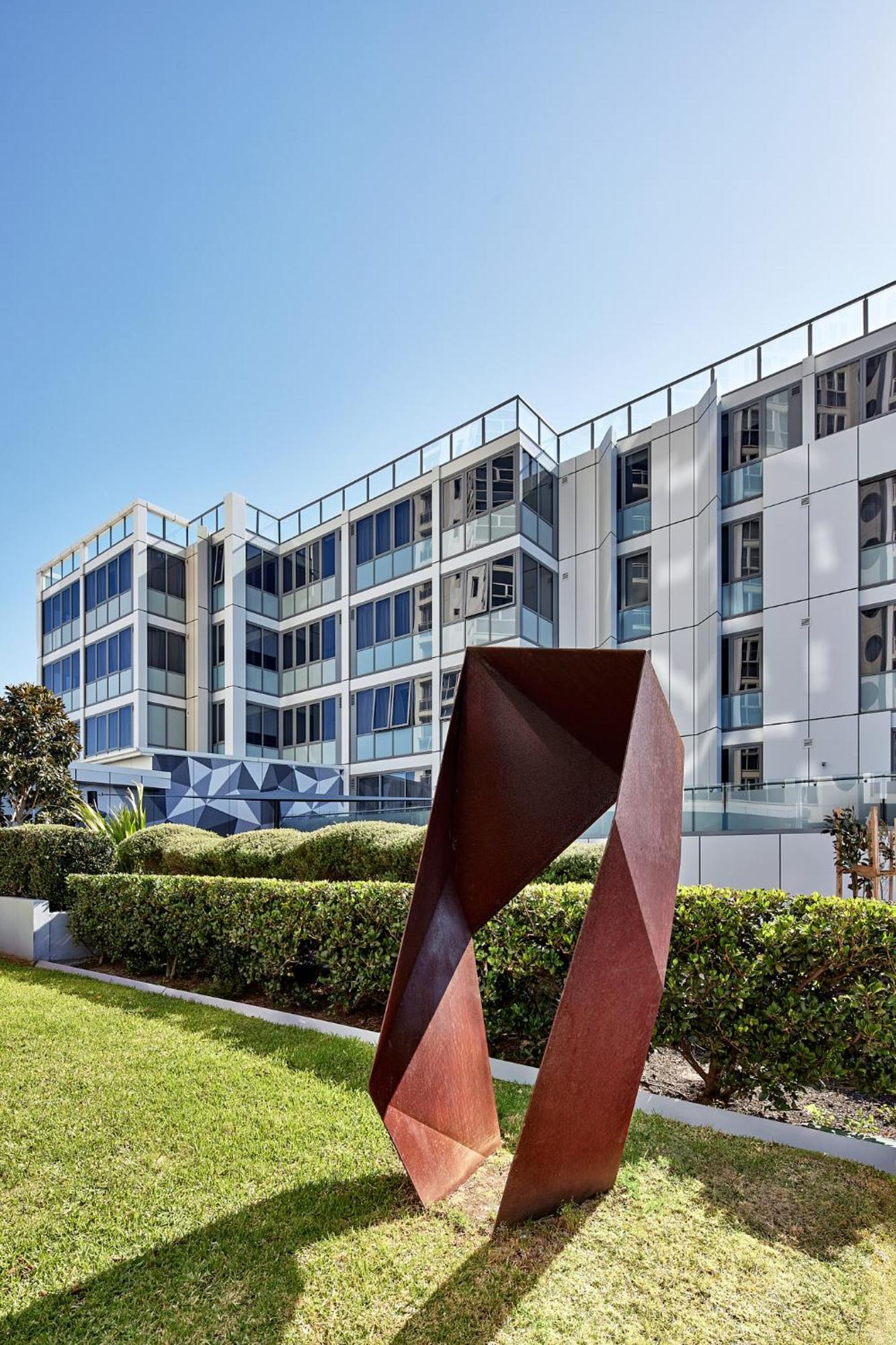 Meriton Suites Bondi Junction Сідней Екстер'єр фото The photo shows a modern building with a sleek white and glass facade, featuring multiple floors and large windows. In the foreground, there is a sculptural piece made of metal, characterized by geometric shapes and a reddish-brown color. The surroun