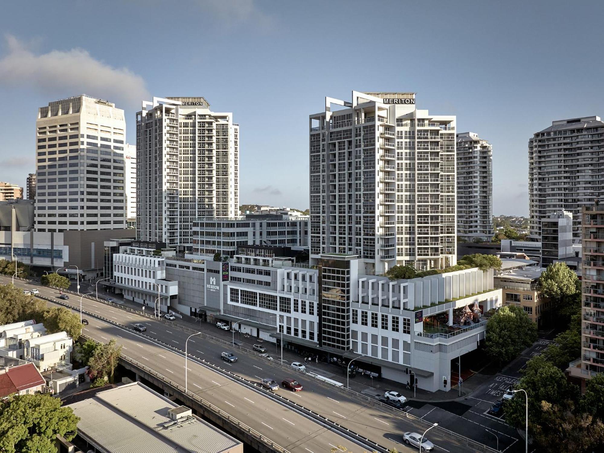 Meriton Suites Bondi Junction Сідней Екстер'єр фото The photo shows a cityscape featuring several tall modern buildings, likely apartments or office complexes. In the foreground, there is a road lined with cars, and the architecture of the buildings varies, with some having sleek glass facades and oth