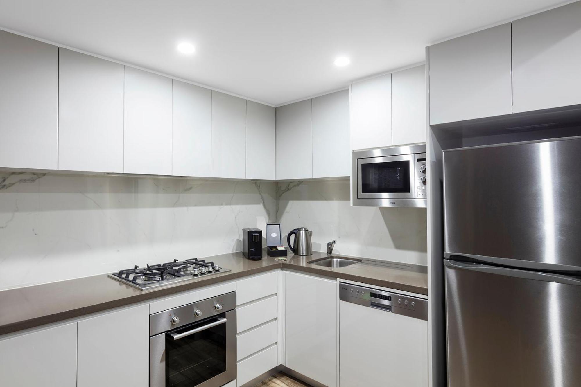 Meriton Suites Bondi Junction Сідней Екстер'єр фото The photo shows a modern kitchen featuring a minimalist design. It has light-colored cabinetry, a countertop, and stainless steel appliances. The appliances include a gas stove with multiple burners, an oven, a microwave, a kettle, and a refrigerator