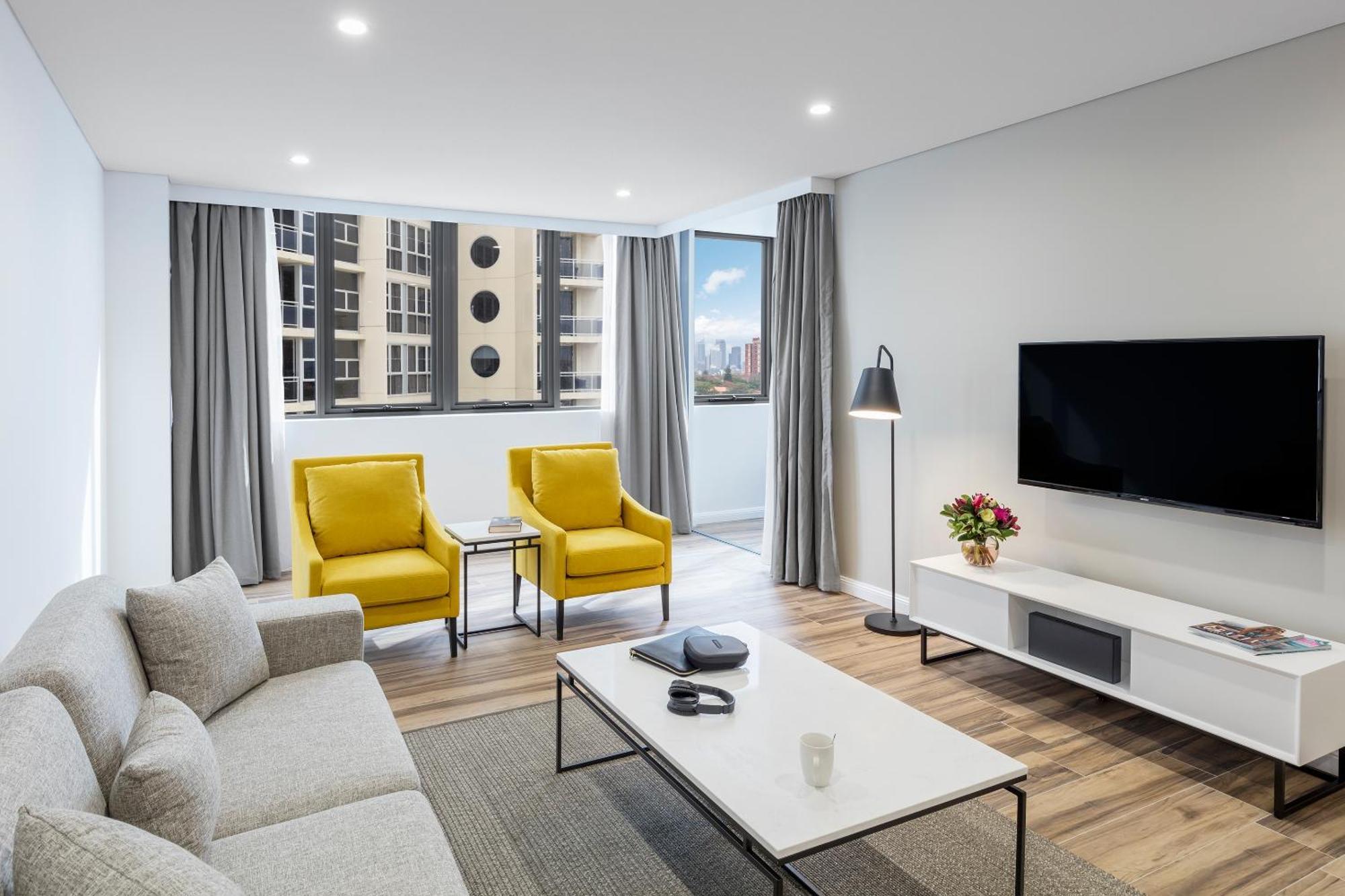 Meriton Suites Bondi Junction Сідней Екстер'єр фото The photo shows a modern, well-lit living room. It features a light gray sofa with a textured fabric. There are two bright yellow armchairs positioned near a window, which allows natural light to enter the space. The room has light gray curtains and 